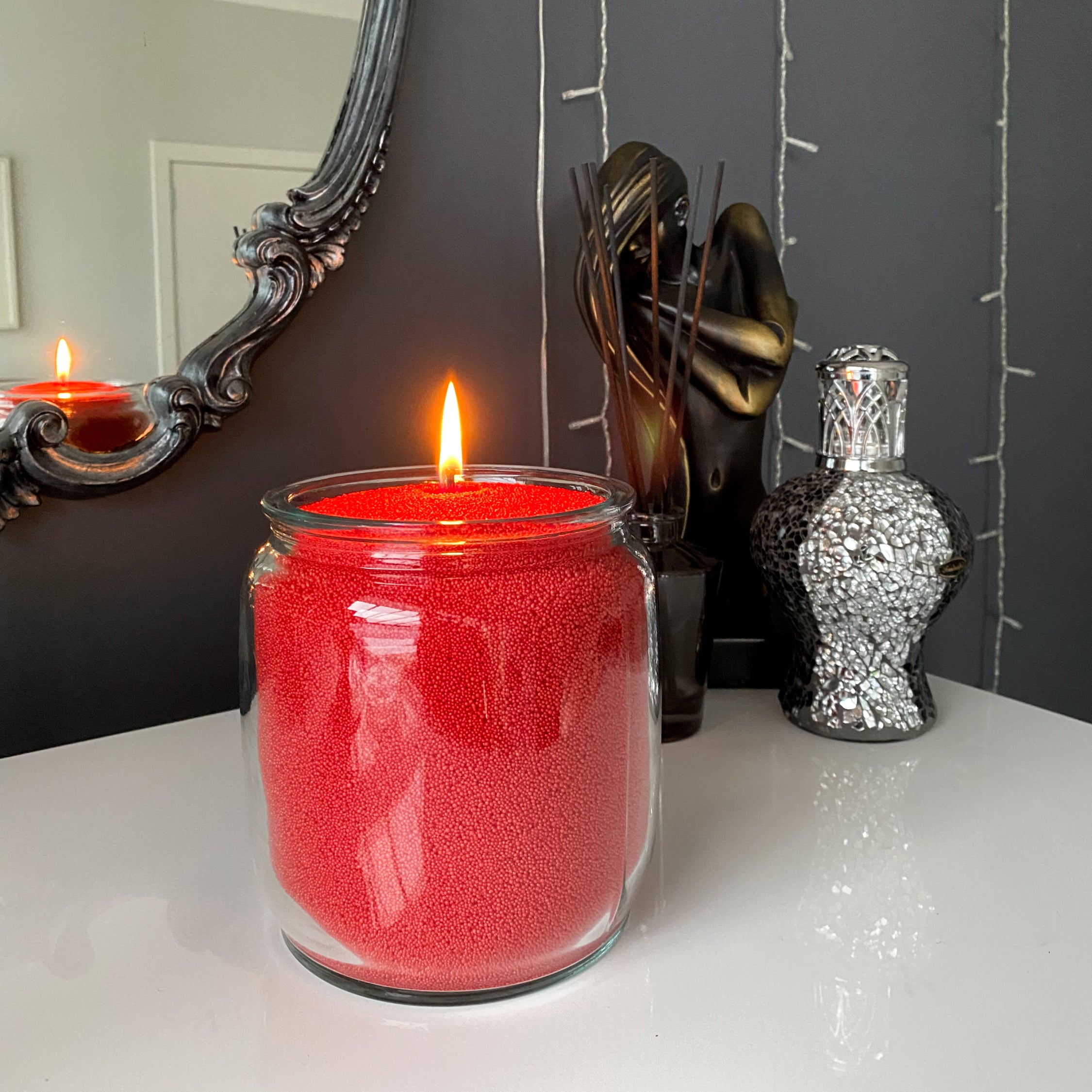 Candle in a glass jar with red sand inside, burning on a surface with decorative items in the background.