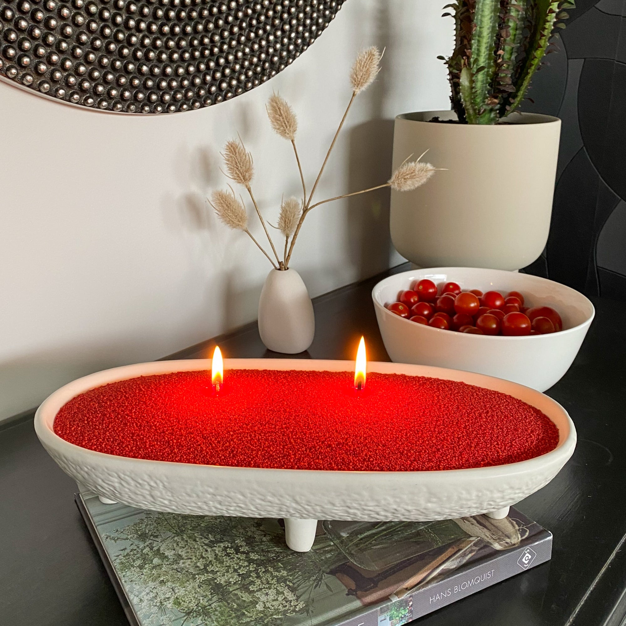 Red sand wax candle in a white holder on a table with decorative items.