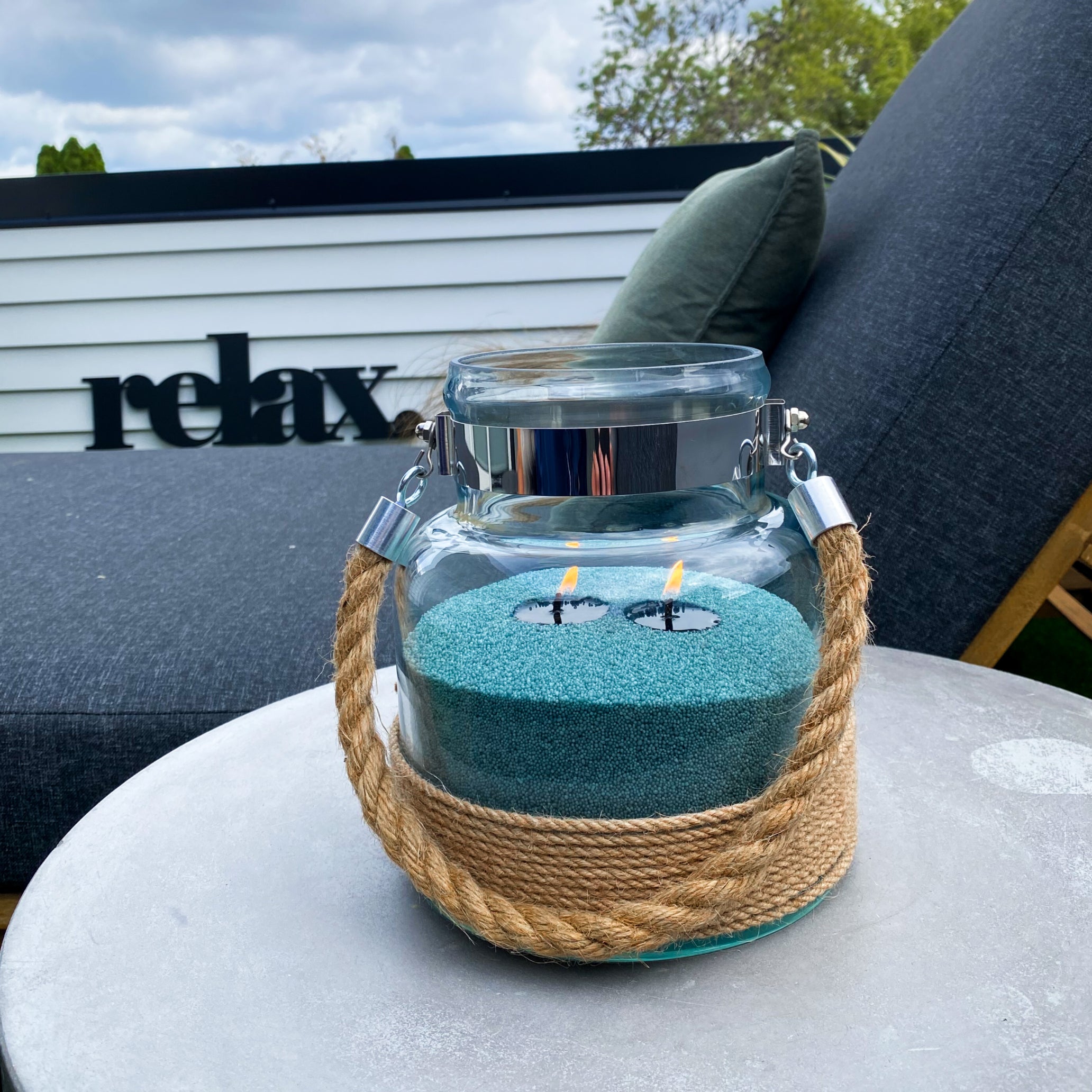 Glass lantern with Dark pine coloured sandwax candle on a table outdoors, with 'relax' sign in the background.